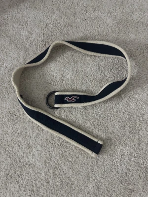 Marinblå och vit tygbälte Hollister - Säljer ett marinblått och vitt tygbälte från Hollister med klassisk logga broderad i rött. Bältet har dubbla D-ringar i metall och är gjort i slitstarkt bomullsmaterial. Perfekt accessoar för att lyfta din outfit.