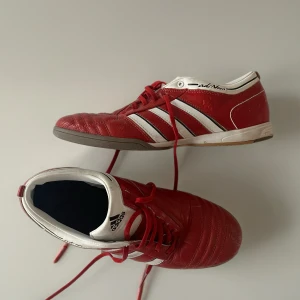 Röda Adidas skor  - Säljer ett par röda Adidas skor med klassiska vita ränder och svart Adidas-logga. Skorna har snörning, låg profil och ovandel i skinn för skön passform. Snygg kontrast mellan rött och vitt, perfekt för dig som letar efter snygga nya skor. 