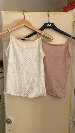 2-pack linnen med rosett och spetskant - Säljer två söta linnen i bomull, ett ljusrosa och ett vitt med små rosa hjärtan. Båda har smala axelband, spetskant upptill och en liten rosett framtill. Perfekta för lager på lager eller till sommaren.