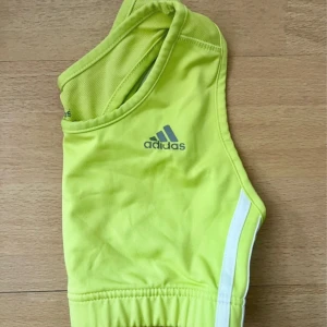 Limegrön adidas sporttopp - Snygg och bekväm sport-BH från Adidas i en fräsch limegrön färg med klassiska vita ränder på sidorna. Sport-bhn är tillverkad i ett mjukt och stretchigt funktionsmaterial som transporterar bort fukt och ger bra stöd vid träning. Perfekt för löpning, gym, dans eller andra träningspass. I mycket fint skick utan fläckar eller skador.  Storlek: M Färg: Limegrön Märke: Adidas  