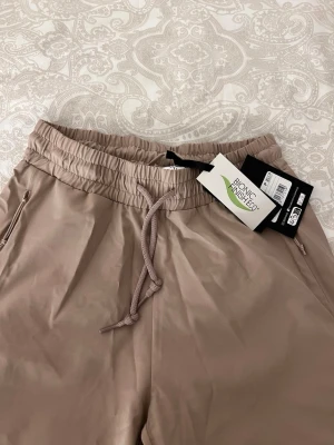 Beige täckbyxor med snörning - Snygga beige täckbyxor med elastisk midja och dragsko för skön passform. Byxorna har dragkedjeförsedda fickor på sidorna och är tillverkade i ett vattenavvisande material. Perfekta för utomhusaktiviteter när du vill hålla dig torr och bekväm.