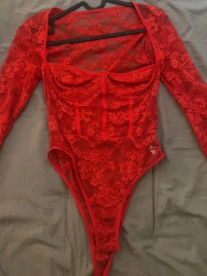 Röd spetsbody med lång ärm - Säljer en röd body i genomskinlig spets med långa ärmar och bygelkupor. Kroppsnära passform och blommigt spetsmönster ger en snygg och trendig look. Perfekt att styla med jeans eller kjol för en cool outfit.
