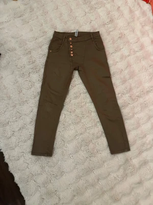 Khaki skinny jeans från 77th Flea - Snygga khaki skinny jeans från 77th Flea med hög midja och fem kopparfärgade knappar framtill. Jeansen har klassiska fickor både fram och bak samt bälteshällor. Materialet är mjukt och stretchigt, perfekt för en trendig och bekväm look.