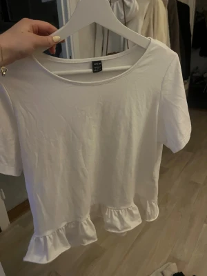 Vit topp med volang från SHEIN - Säljer en vit topp från SHEIN med rund halsringning och korta ärmar. Toppen har en söt volangdetalj längst ner som ger en extra touch. Perfekt till jeans eller kjol för en enkel men gullig look.