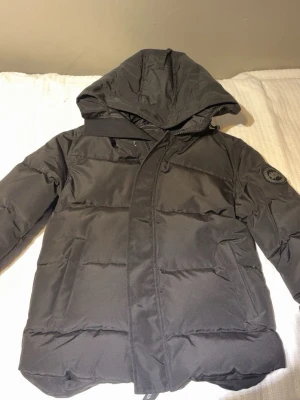 Svart pufferjacka från Canada Goose - Säljer en svart pufferjacka från Canada Goose med huva och diskret logga på ärmen. Jackan har dragkedja och stora fickor framtill. Perfekt för kalla dagar och riktigt snygg till streetwear-stilen. Helt ny!!!