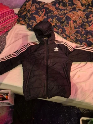 Svart Adidas pufferjacka med huva - Säljer en svart pufferjacka från Adidas med klassiska vita ränder på ärmarna och Adidas-logga på bröstet. Säljer den för att jag nu har växt ur den. Jackan har huva, dragkedja framtill och quiltad design. Perfekt för kyligare dagar och riktigt snygg streetstil. Kan tvättas in den skickas