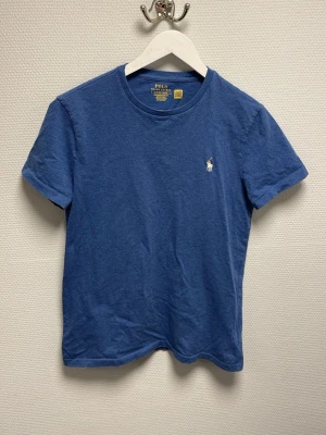 Blå t-shirt från Polo Ralph Lauren - Blå t-shirt från Polo Ralph Lauren i custom slim fit. Klassisk design med korta ärmar, rund halsringning och den ikoniska broderade loggan på bröstet. Tillverkad i mjuk bomull som känns skön mot huden. Perfekt för en clean och stilren look. Ny pris 900-1000kr.