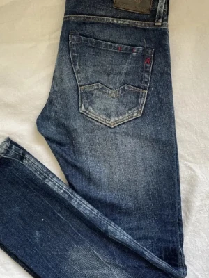 Replay Anbass blå jeans, storlek 29 - Säljer ett par Replay Anbass jeans i klassisk blå tvätt med snygga slitningar och kontrastsömmar. Modellen har normal passform och raka ben, med detaljer som Replay-märke på fickan och röd söm på insidan. Perfekta för dig som gillar en tidlös jeanslook.