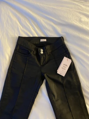 Svarta coated byxor från Nelly - Snygga svarta coated byxor från Nelly med låg midja och utsvängda ben. Byxorna har dubbla silverknappar framtill, klassiska fickor och en cool broderad detalj på bakfickan. Helt nya, endast testade. Säljer pga för små!