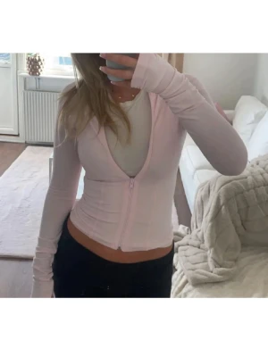 Ljusrosa croppad kofta med dragkedja - Säljer en ljusrosa croppad kofta med dragkedja framtill. Tröjan har lång ärm och är figurnära i modellen, perfekt för lager på lager eller som ett statement-plagg. Enkel och stilren design som passar till mycket.