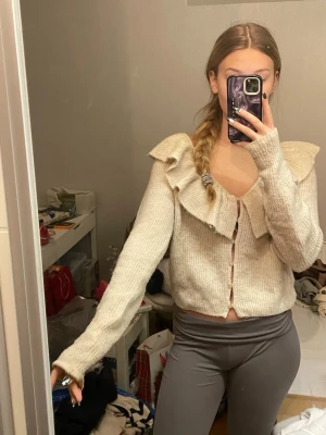 Beige stickad kofta med volang - Superfin beige stickad kofta med volangdetalj runt hela axelpartiet och knäppning framtill. Koftan är croppad och har långa ärmar, perfekt att styla med jeans eller kjol för en söt look.