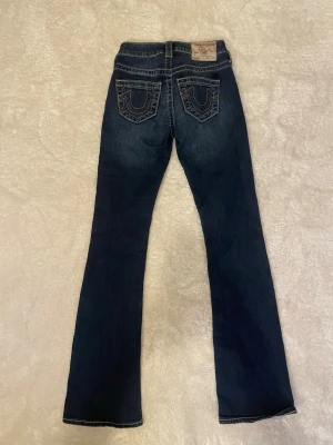 True Religion blå bootcut jeans - Säljer ett par mörkblå jeans från True Religion med kontrastsömmar och ikoniska bakfickor. Modellen är bootcut med låg midja. Det står på lappen i byxorna att de är mid waist men jag upplever dem mer low waist! Sparsamt använda och har ej några tecken på ”skador”💓 Dessa byxor är perfekta för en stilren och ikonisk look!🙏🏼💓 Köpare står för frakt💓