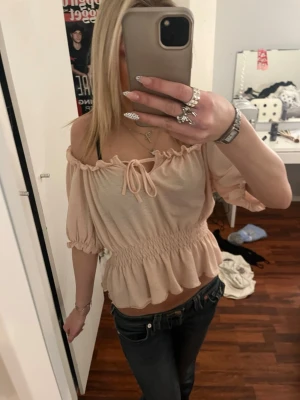 Beige offshoulder blus med volang - Supersöt beige offshoulder blus med volangkant och smock i midjan. Toppen har korta puffärmar och en söt knytning framtill vid halsen. Perfekt för en somrig look och passar till både jeans och kjol.