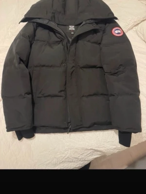 Svart dunjacka från Canada Goose - Säljer en svart dunjacka från Canada Goose i storlek L men passar M. Jackan har en klassisk puffer-look med huva, ribbade muddar och dragkedja framtill. Perfekt för kalla dagar och riktigt snygg med den ikoniska loggan på ärmen.