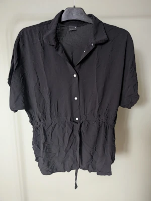 Svart blus med knyt från Gina Tricot - Svart blus från Gina Tricot i luftig bomull med korta ärmar, knappar framtill och knyt i midjan. Skjortan har en avslappnad passform och klassisk krage, perfekt för en chill och enkel stil.