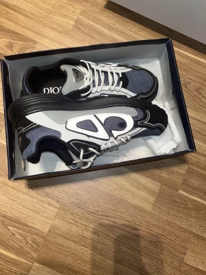 Dior sneakers i svart, grå och vit - Säljer ett par feta Dior sneakers med svart, grå och vit färgkombination. Skorna har reflekterande detaljer, meshpaneler och vita snören. Tjock sula och sportig vibe, perfekta för dig som vill sticka ut. Märket syns tydligt på plösen och insidan.