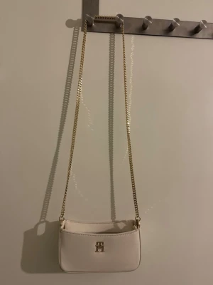 Vit axelväska från Tommy Hilfiger - Säljer en stilren vit axelväska från Tommy Hilfiger med guldfärgad kedjerem och TH-logga framtill sopm detalj. Väskan är i slätt material och har dragkedja upptill. Perfekt storlek för det viktigaste och ger en clean look till din outfit.