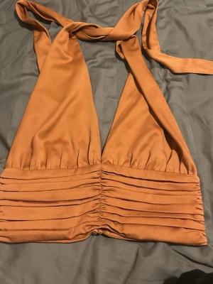 Halterneck korsettopp i satin-look - Säljer en snygg halterneck korsettopp i varm orange satin-look. Toppen har breda knytband i nacken, djup v-ringning och dekorativa knappar framtill. Midjan är rynkad och ger en cool, croppad siluett. Perfekt för dig som vill sticka ut lite extra!
