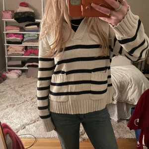 Randig stickad tröja Holly & Whyte - Säljer en krämvit och marinblå randig stickad tröja från Holly & Whyte. Tröjan har v-ringad krage, två fickor framtill och bred ribbad mudd vid ärmslut och nederkant. Perfekt till jeans för en chill look.