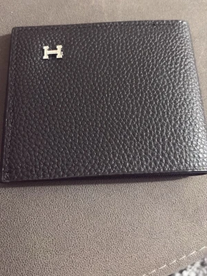 Svart plånbok från h  i skinn - Snygg svart plånbok från H  i exklusivt präglat skinn. Klassisk design med silverfärgad H-detalj på framsidan och flera kortfack på insidan. Perfekt för dig som vill ha en stilren och lyxig accessoar i fickan.