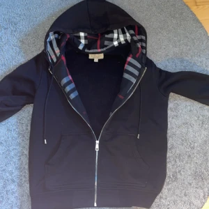 Svart Burberry hoodie med dragkedja - Svart hoodie från Burberry med klassiskt rutigt foder i huvan och silverfärgad dragkedja. Tröjan har snörning i huvan, känguruficka och är tillverkad i mjuk bomull. Perfekt för dig som vill ha en stilren och lyxig streetwear-look.Är lite solblekt i luvan men syns knappt 