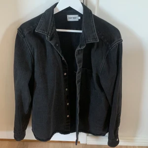 Svart overshirt från East West - Svart overshirt från East West i 100% bomull. Klassisk skjortkrage, knäppning framtill och en bröstficka. Snygg loose fit och passar perfekt som lager över t-shirt eller hoodie. Enkel och stilren design med långa ärmar och diskreta sömmar.