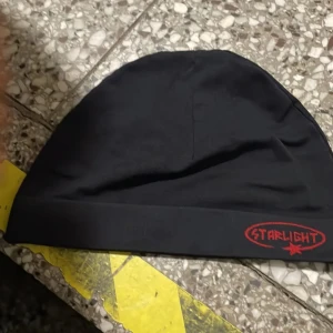 Svart mössa från Starlight - Svart, enkel och stilren mössa från Starlight med röd broderad logga framtill. Mössan har en klassisk form och är tillverkad i mjukt material som känns skönt mot huden. Perfekt för dig som gillar minimalistisk streetstyle.