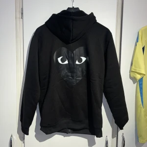 Svart hoodie med hjärta och ögon - Svart hoodie med dragkedja och huva, prydd med ett stort svart hjärta med ögon på ryggen och ett mindre likadant tryck på bröstet. Tröjan har kängurufickor och snörning i huvan. Perfekt för dig som gillar streetstyle och unika detaljer.
