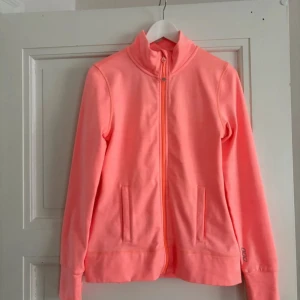 Rosa träningsjacka från Lorna Jane - Sportig och snygg rosa träningsjacka från Lorna Jane. Jackan har hel dragkedja framtill, höga fickor med dragkedja och stårkrage. Tillverkad i mjuk polyester, perfekt för aktiva dagar. Liten logga på ärmen och i nacken.