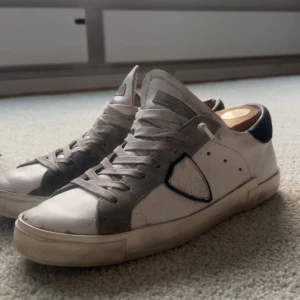 Philippe Model vita och grå sneakers - Snygga Philippe Model sneakers i vitt och grått med svarta detaljer. Skorna har rund tå, platt sula och snörning. Ovandelen är i skinn och mocka, med en ikonisk sköld på sidan. Perfekta för dig som gillar stilrena och exklusiva sneakers.                 Om dustbag och kartong önskas är de 175kr extra, de är bra skick, om man vill ha de mer rena är det bara säga till, kan mötas upp eventuellt posta de. 