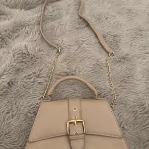 Beige axelväska med gulddetaljer - Stilren beige axelväska med guldfärgad kedja och spänne framtill. Väskan har ett topphandtag och justerbar axelrem i matchande färg. Snygg och enkel design som passar till många outfits. Materialet är slätt och ger ett lyxigt intryck.