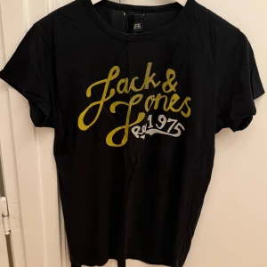 Svart Jack & Jones t-shirt med tryck - Svart t-shirt från Jack & Jones med stort gult och vitt tryck på bröstet där det står 'Jack & Jones Est. 1975'. Klassisk rund hals och korta ärmar. Skön passform och tillverkad i mjuk bomull. Storlek M.