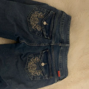 Blå jeans med broderade bakfickor - Snygga blå jeans med unika broderade detaljer på bakfickorna och dubbla knappar framtill. Klassisk femficksmodell i jeansmaterial, perfekt för dig som vill sticka ut lite extra. Jeansen har en rak passform och låg midja.