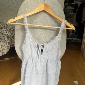 Ljusblå linnetopp från Gina Tricot - Supersöt ljusblå linnetopp från Gina Tricot med spetsdetaljer längs axelbanden och en liten knytning framtill. Toppen har en lätt rynkad struktur och är ärmlös, perfekt för varma dagar. Luftig och feminin design som ger en chill vibe.