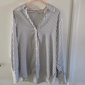 Randig vit skjorta från H&M, stl L - Vit skjorta från H&M med svarta vertikala ränder och klassisk krage. Skjortan har långa ärmar med breda manschetter och knäppning framtill. Luftig och loose fit, perfekt för en chill och stilren look.