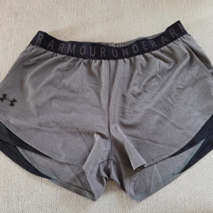 Grå träningsshorts från Under Armour - Säljer ett par grå träningsshorts från Under Armour med svart resår i midjan där loggan syns tydligt samt fickor. Shortsen är i loose fit och tillverkade i ett lätt syntetmaterial, perfekta för träning. Klassisk sportig look med diskret logga på benet.