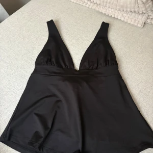 Svart omlottopp med rosett från Nelly - Snygg svart omlottopp från Nelly med djup v-ringning och breda axelband. Toppen har en knytbar rosett i ryggen som ger en extra cool detalj. Materialet är mjukt och har en lätt glansig finish som känns lyxig. Perfekt att styla med jeans eller kjol.