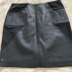 NySvart skinnkjol från River Island - Snygg svart kortkjol från River Island i skinnimitation med mockadetaljer på sidorna. Kjolen har en markerad dragkedja bak i guld och en cool geometrisk design. Perfekt för dig som gillar en edgy och trendig look. Oanvänd  36 Stl 