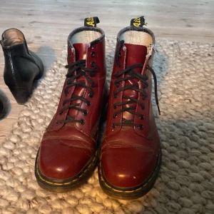Vinröda Dr. Martens boots i skinn - Säljer ett par klassiska vinröda Dr. Martens boots i skinn med svarta snören och ikonisk gul söm runt sulan. Storlek 39-40 SUPER BRA PRIS. Skorna har rund tå, platt sula och den typiska Dr. Martens-dragfliken baktill. Perfekta för dig som vill ha en edgy och tidlös stil. Ganska slitna. Hör av dig om du har frågor eller funderingar!
