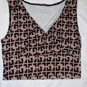 Omlottopp - Säljer en beige och svart omlottopp med grafiskt mönster. Toppen är ärmlös och har en snygg v-ringning framtill. Materialet känns mjukt och stretchigt, perfekt för att skapa en cool look. Passar dig som gillar unika mönster och vill sticka ut.
