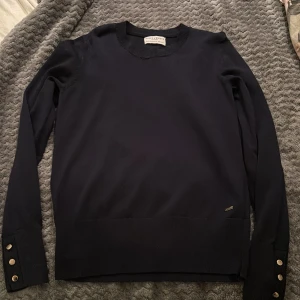 Mörkblå tröja Holly & Whyte S - Mörkblå långärmad tröja från Holly & Whyte med rund halsringning och snygga guldfärgade knappar vid ärmsluten. Tröjan är stickad och har en liten metallbricka med logga nedtill. Perfekt för dig som gillar stilrena detaljer.