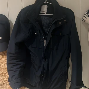 Fieldjacka från J.Lindeberg - Snygg mörkblå vindjacka från J.Lindeberg med hög krage, dragkedja och knappar framtill. Jackan har flera fickor med lock och tryckknappar samt justerbara detaljer vid ärmslut och midja. Storlek M. Det nya priset är 6000 kr på jacka