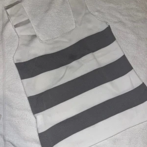 Topp - Säljer en stilren ärmlös topp med breda grå och vita ränder. Toppen har fyrkantig ringning och är tillverkad i ett mjukt, stickat material. Perfekt för en chill och trendig look. Aldrig använd