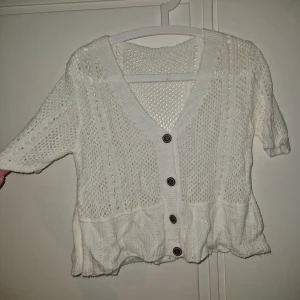 Vit stickad kofta med knappar - Superfin vit stickad kofta med v-ringning och bruna knappar framtill. Koftan har korta ärmar, ett luftigt hålmönster och en volangliknande kant nedtill. Perfekt att slänga över en topp för en söt och chill look.