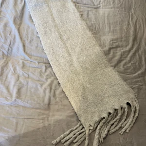 Ljusgrå ullhalsduk med fransar - En stilren ljusgrå halsduk i mjuk ull med klassiska tvinnade fransar i ändarna. Halsduken är rektangulär och har en enkel, enfärgad design som passar perfekt för kyliga dagar. Perfekt accessoar för att hålla värmen och lyfta din outfit.