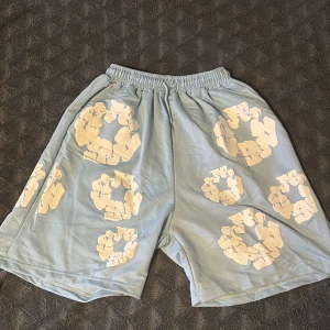 Blå shorts med blommigt tryck från Denim Tears - Säljer ett par ljusblå shorts från Denim Tears med elastisk midja och vita blommiga tryck över hela plagget. De har en loose passform och är tillverkade i mjuk bomull, perfekt för chill dagar. Bakficka på ena sidan för extra stilpoäng.