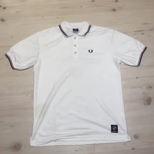 Vit pikétröja från Fred Perry - Klassisk vit pikétröja från Fred Perry med broderad logga på bröstet. Tröjan har korta ärmar, krage med knappar och tunna marinblå och röda ränder på krage och ärmslut. Perfekt för en clean och sportig stil.