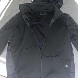 Svart pufferjacka från Balmain strl 48 - Snygg svart pufferjacka från Balmain med stora logotyper på ärmar och huva. Jackan har quiltad baksida, dragkedja framtill och två stora fickor. Materialmix med glansig polyester på ryggen och mjukare tyg på ärmar och framsida. Perfekt för dig som vill sticka ut. Du kan även välja att ta bort luvan då den kan gå in i jackan.