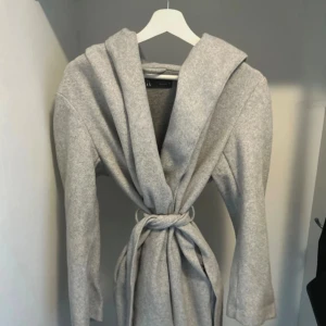 Ljusgrå kappa med huva och bälte - Snygg ljusgrå kappa från H&M med stor huva och knytskärp i midjan. Jackan har lång ärm och en rak, avslappnad passform. Perfekt för dig som gillar stilrena och mysiga ytterplagg.