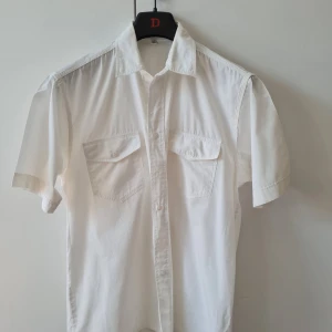 Kortärmad skjorta med fickor strl S (M) (herr) - Vit skjorta i 100% bomull med korta ärmar och två bröstfickor med lock. Klassisk krage och knäppning framtill. Känns stor i storleken. Bröstmått 114cm. Storlek S (känns som M). Kan hämtas i Vallentuna.